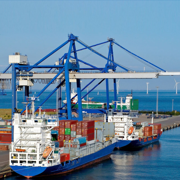 Zwei mittelgroße Handelsschiffe liegen am Kai. Beide sind mit Containern beladen. In der oberen Bildhälfte sind die riesigen Kräen vor blauem Himmel zu sehen, im Hintergrund ist das offene Meer.