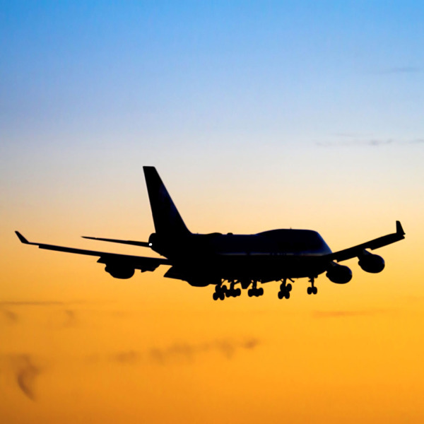 A plane is flying into the dusk. It's the silhouette of a Boeing 747. The sky in the upper half is light blue, in the lower half light orange.