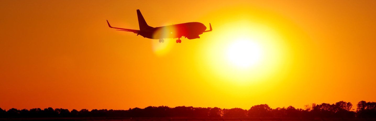 Ein Flugzeug startet in die Dämmerung und ist bereits in der Luft. Im Bild zentral ist die Sonne, die direkt in die Kamera scheint. Das Flugzeug ist als Schattenriss zu erkennen, das Bild ist komplett in den verschiedensten Orangetönen.