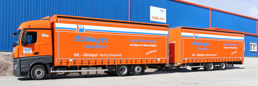 A gigaliner, which is an extra long truck & trailer combination in it's company colors orange and blue is located in front of a blue and orange warehouse.
