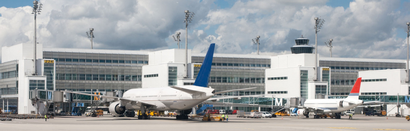 One can see the buildings of a new, modern airport. In front of it two passenger planes are parked, and you can see different units as well as cars. In the background you can see the airport tower.