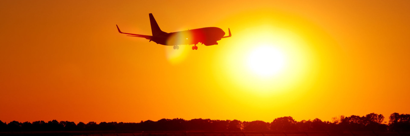 An airplane takes off in the direction of the sun. The sun is about to set and is shining directly into the camera. The sky is a mix of many orange tones.