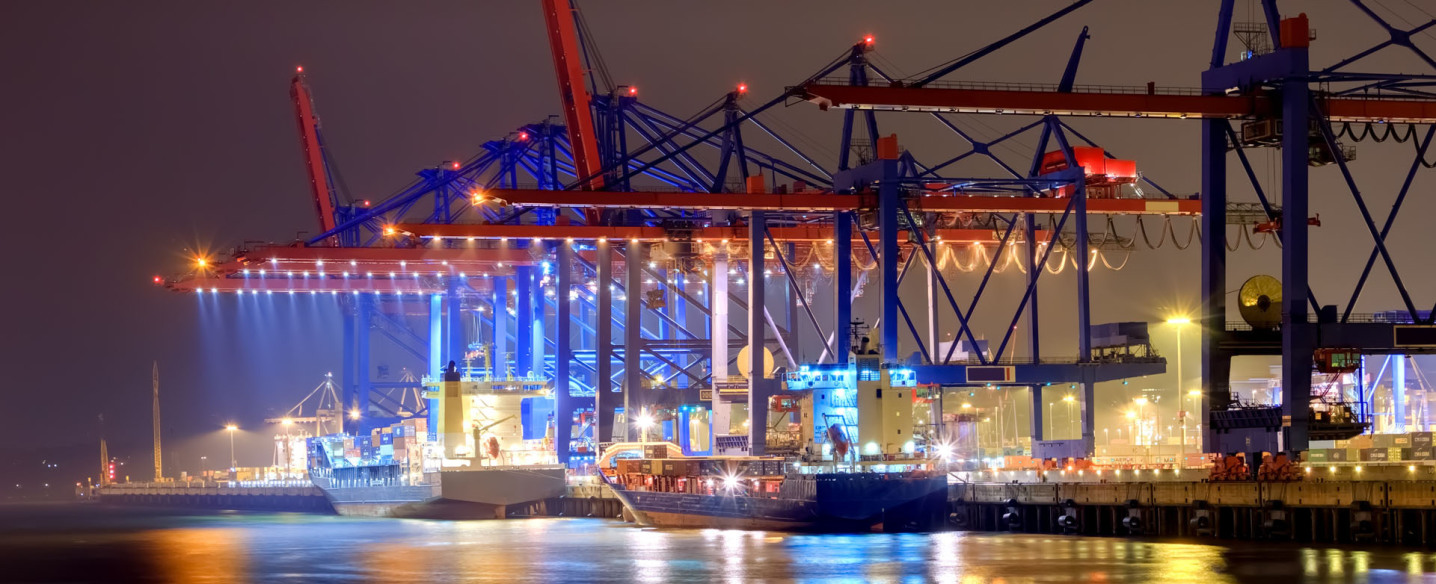 Night in a very modern cargo port. Two ships are docked at the quay. In the upper half of the picture is full of crane equipment in the colors blue and orange. In the foreground you can see water and the mirror of the subject above.