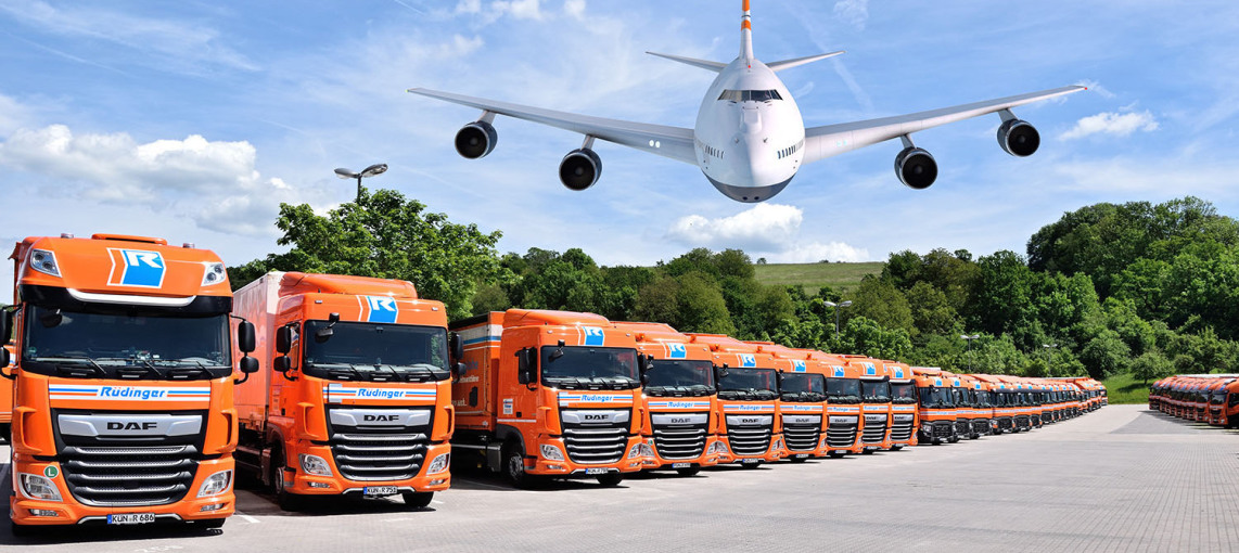It's a photo composition: You see two long rows of parked trucks with trailers down to the depth of the picture. Behind them are trees. In the sky, an airplane is heading low toward the viewer.