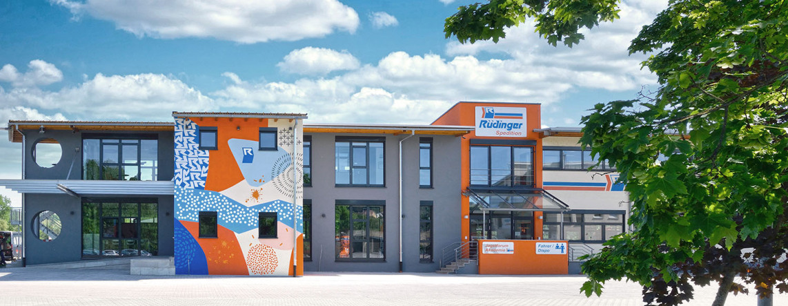 The administrative building of the freight forwarding company is a work of art: The employees designed it in the colors orange and blue together with an artist. Two other parts are gray, one orange. The sky is blue and there are white clouds.
