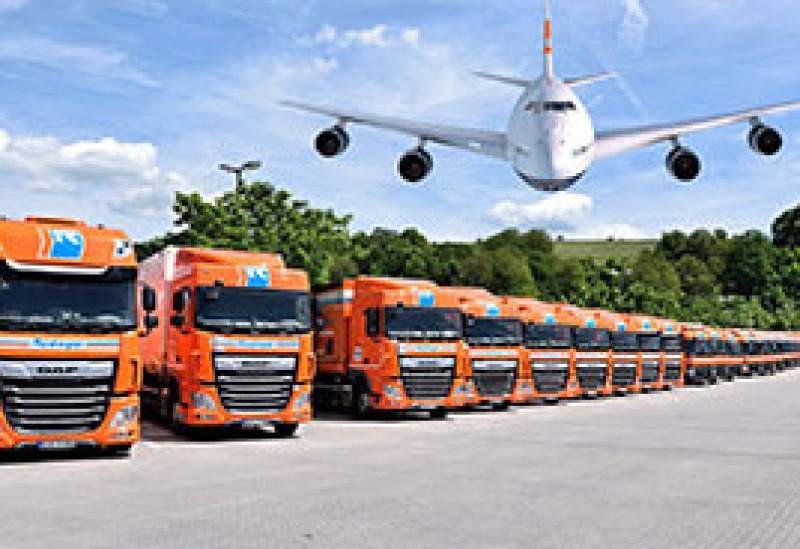 Vom linken bis zum rechten Bildrand steht eine riesige Flotte an orangenen Planentiefladern. Darüber fliegt eine Boeing 747 im Tiefflug auf den Betrachter zu. Es ist eine Fotomontage.