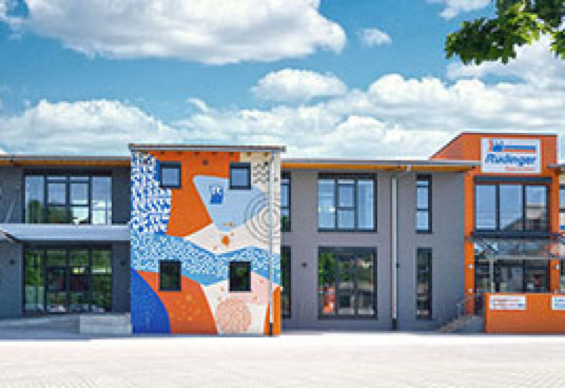 Das linke Drittel des Speditions-Neubaus: das Jagstforum. Ein Drittel ist ein riesiges Fassaden-Kunstwerk in Blau und Orange. Der Himmel ist blau, darin sind weiße Schönwetterwolken.
