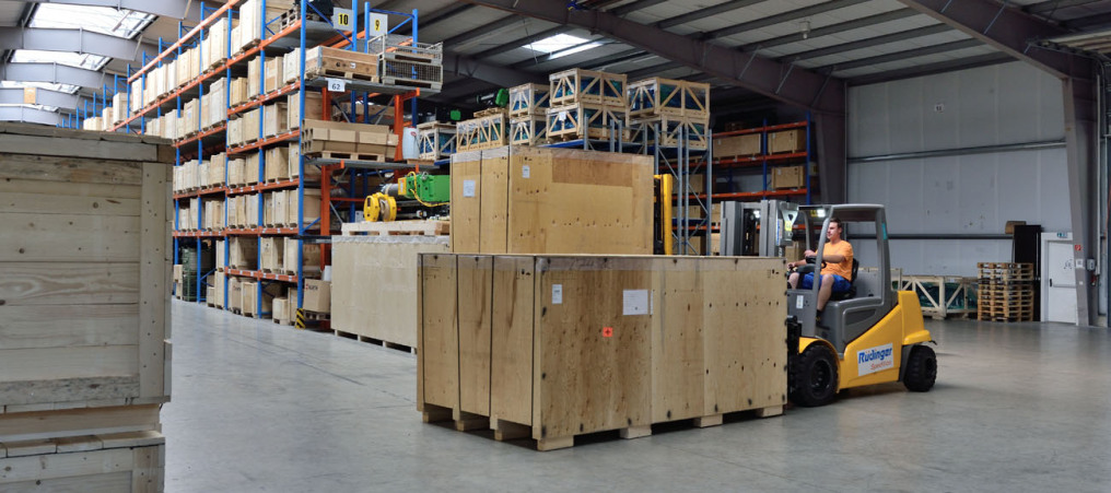You look into a modern warehouse, the levels are full of goods. In front you can see a forklift that has loaded a large wooden crate.