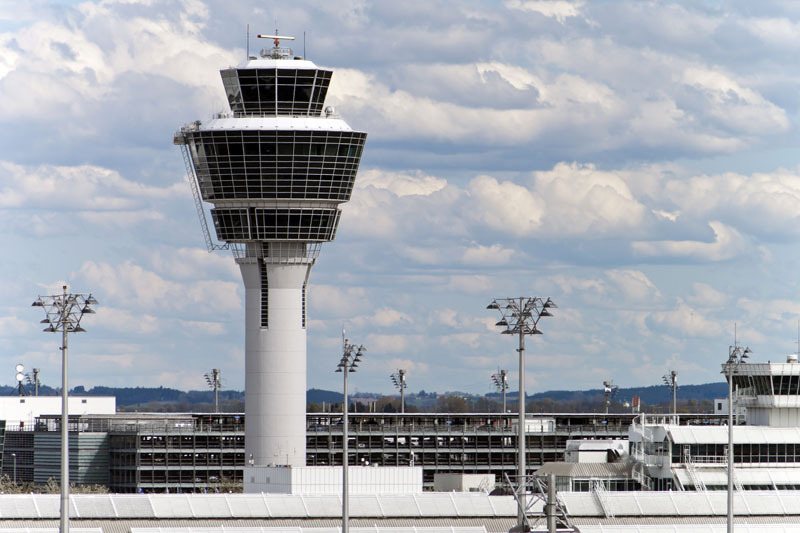 On a modern airport it is a top modern airport tower. In the lower part of the photo are more modern buildings and light installations. Above the sky is covered with many clouds.