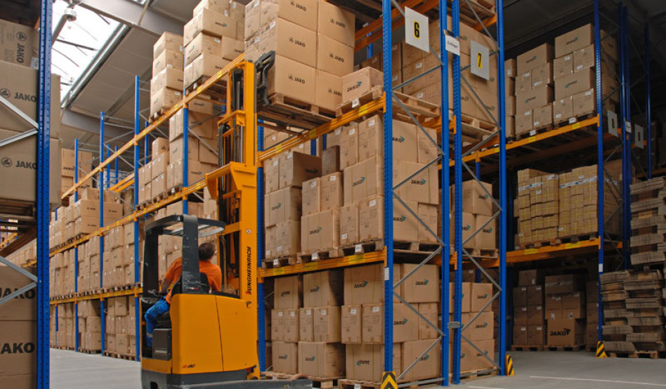 In a newly built warehouse you can see many shelves in the colors blue and orange. All areas are filled with goods. In the foreground an employee in a forklift lifts a pallet to the third level.