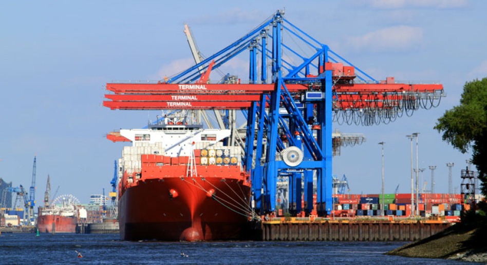 In the center of the picture you can see a huge container ship centrally from the front, to the left of it a crane system in the colors blue and orange. On the left you can see another ship at the quay in the distance, on the right there are containers.