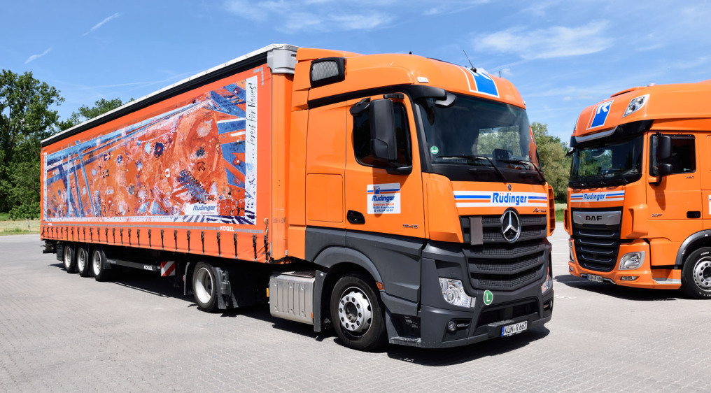 It is a special tarp low loader truck and trailer over almost the whole picture. The tarp is decorated with artwork in blue and orange colors. On the right you can see the front half of a second truck.