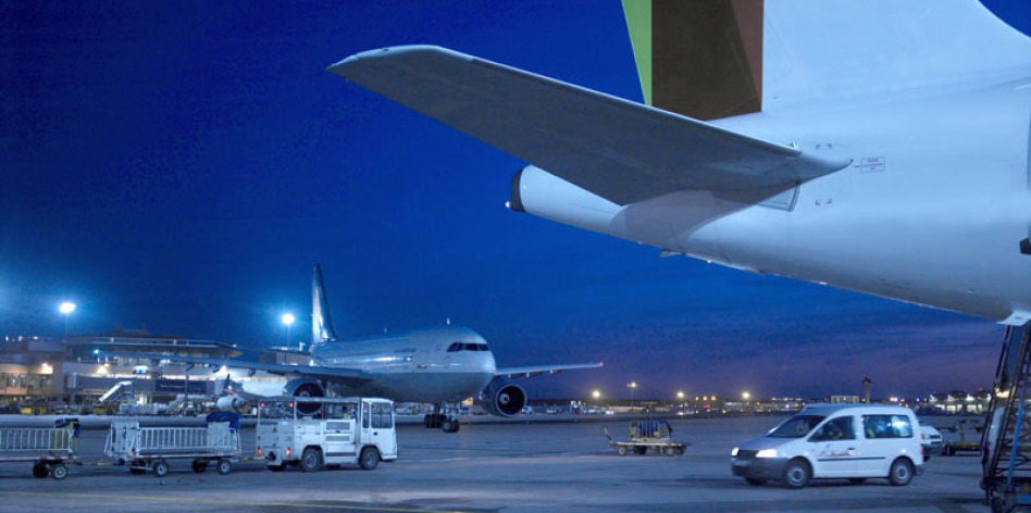 Es ist Nacht auf einem Flughafen, dort wo Flugzeuge geparkt sind. In der linken Bildhälfte sieht man einen Jet komplett, in der rechten ist es das hintere Stück, dafür riesig. Im Bild fahren mehrere Fahrzeuge herum. Es ist in der Blauen Stunde fotogarfier