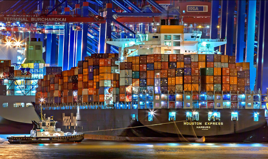 Eine Nachtaufnahme zeigt ein supergroßes Containerschiff mit huderten bunter Container am Kai festgemacht. Im Vordergurnd schwimmt ein Schlepper, im Hintergrund sieht man die beleuchteten Kräne.