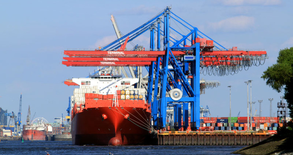 Ein großes Containerschiff ist am Kai im Hafen festgemacht. Man sieht von vorne auf das Schiff. Ebenfalls in der Bildmitte ist eine Kraneinheit, rechts sieht man Container an Land. Links kann man weitere Schiffe erkennen.